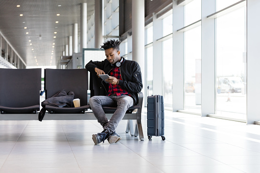 African guy waiting at airport lounge using his digital tablet. Young man sitting at airport terminal bench with tablet pc.