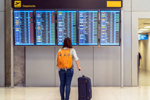 Back side of traveler with luggage standing over the flight board for check-in at the flight information screen in modern an airport, travel and transportation with technology concept. stock photo