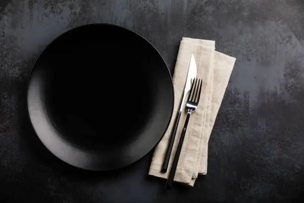 Table setting with empty black plate, fork and knife cutlery and linen napkin on dark table