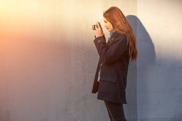 photographe féminin souriant dans la veste restant devant le mur prêt à faire la nouvelle photo. fusée de soleil - concrete wall flash photos et images de collection