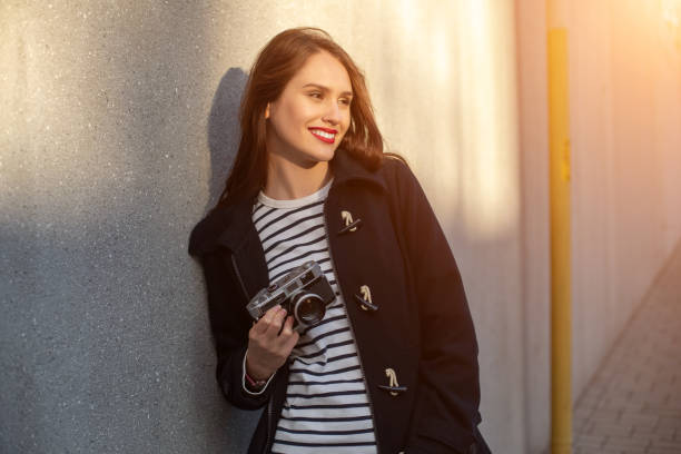 photographe féminin souriant dans la veste restant devant le mur prêt à faire la nouvelle photo. fusée de soleil - concrete wall flash photos et images de collection