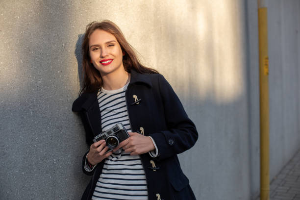 photographe féminin souriant dans la veste restant devant le mur prêt à faire la nouvelle photo. fusée de soleil - concrete wall flash photos et images de collection