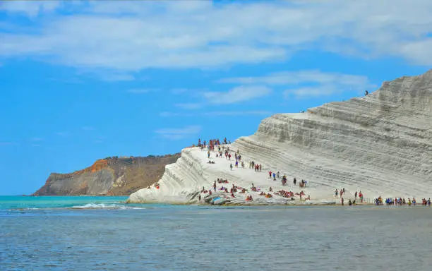 white cliffs naturally made of smooth pug at Scala dei Turchi beach full of people with turquoise mediterranean sea and blue cloudy summer sky near Agrigento, Sicily, Italy