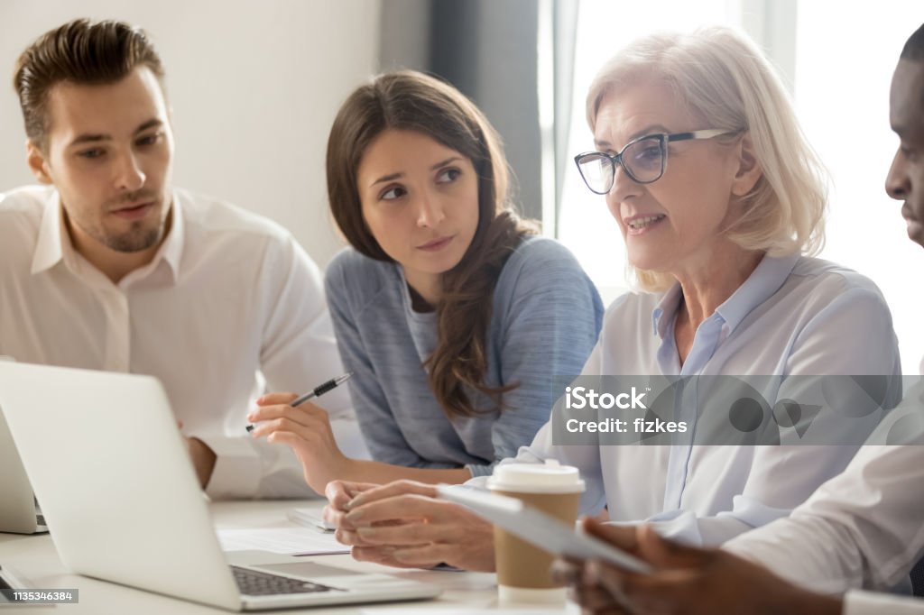 Focused young interns making notes listening to old female manager Focused young students interns workers making notes listening to old senior aged female manager coach mentor leader teacher talking at group office meeting instructing business work team with laptop Family Stock Photo