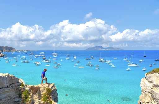 02.09.2018. Man on the top of the rock cliff looks at paradise clear torquoise blue water with boats and cloudy blue sky in background in Favignana island, Cala Rossa Beach, Sicily South Italy.
