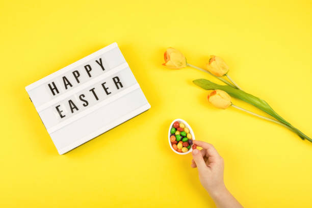 istock Easter composition, greeting card with child hand, lightbox text Happy Easter, yellow tulips, chocolate eggs with multicolored dragee on color background 1135349176