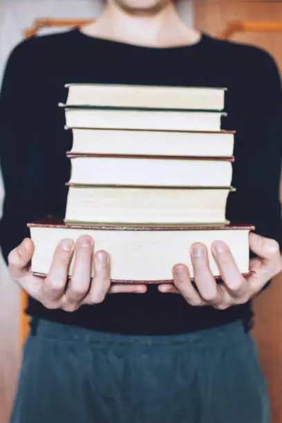 Photo of Man, Student holding many books in hands on the background of bookshelves. Male hands hold a large stack of books.