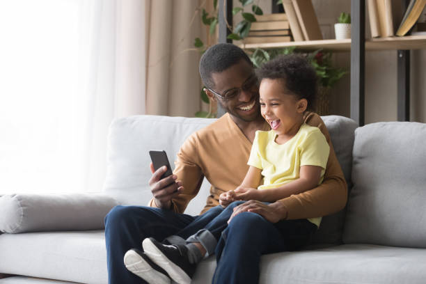 feliz padre africano y niño hijo riendo mirando en el teléfono móvil - mirar un objeto fotos fotografías e imágenes de stock