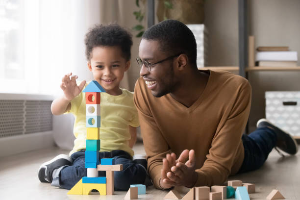 Happy little son playing with black dad using wooden blocks Happy cute little son playing game with black dad baby sitter building constructor tower from multicolored wooden blocks, african family father and toddler child boy having fun on warm floor at home genderblend stock pictures, royalty-free photos & images