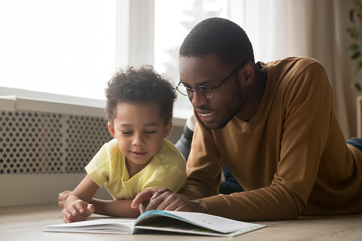 Happy black father baby sitter and toddler son having fun with book lying on warm floor, african dad teaching little kid boy learning to read, parent with children educational activities at home