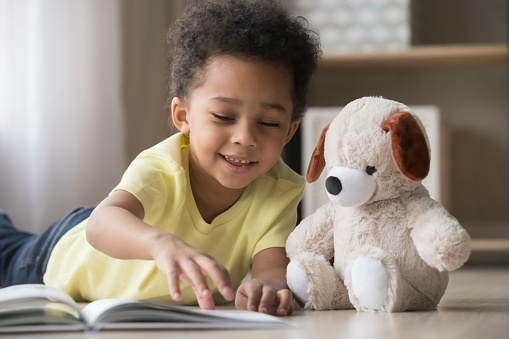 Happy mixed race little boy enjoying playing alone reading book to fluffy toy lying on warm floor, cute smiling african american kid having fun at home, creative child activity, underfloor heating
