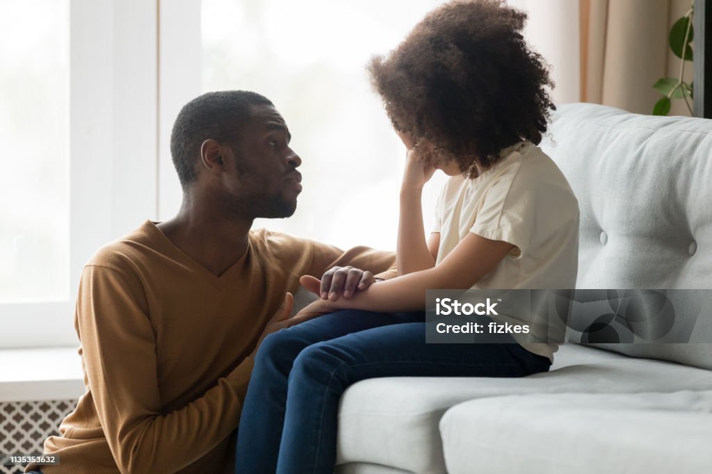 Loving african dad comforting crying kid daughter showing empathy Loving african american dad comforting crying sad kid daughter holding hand supporting little stressed school girl in tears, black father consoling talking to upset child giving empathy protection Child Stock Photo