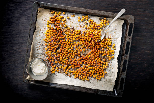 Tray of roasted or baked seasoned chickpeas. Tray of baked or roasted chick-peas seasoned with paprika and oregano. Overhead view looking down, photographed on a baking tray with baking parchment on it, colour, horizontal with some copy space. With a side bowl of soured cream or creme fraiche. chick pea photos stock pictures, royalty-free photos & images