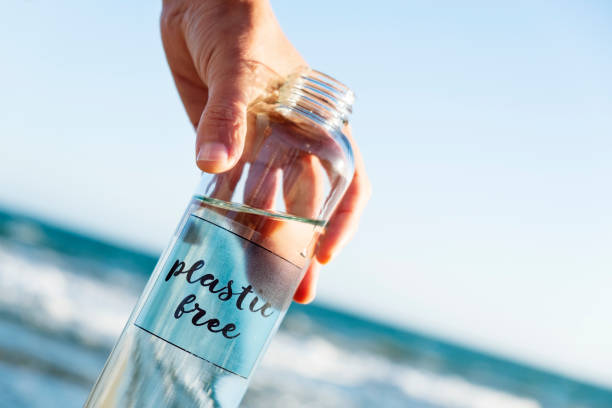 reusable water bottle with the text plastic free closeup of a caucasian man holding a glass reusable water bottle with the text plastic free written in it, on the beach, with the ocean in the background plastic free photos stock pictures, royalty-free photos & images