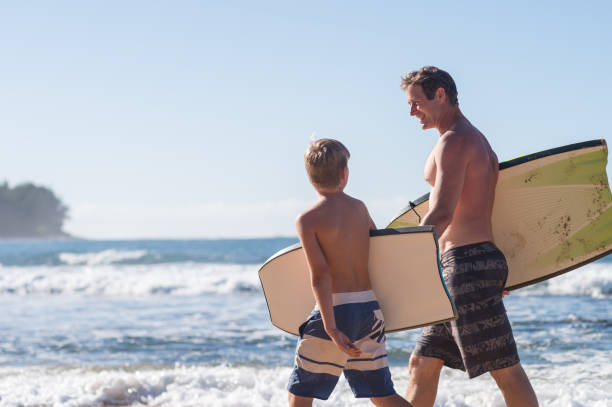 Dad giving his son a boogie boarding lesson A middle-aged dad walks with his son along the sandy beach. They're carrying boogie boards and preparing to hop in the ocean for an afternoon of fun! body board stock pictures, royalty-free photos & images