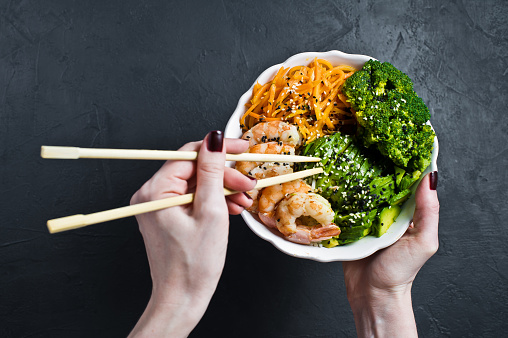 Girl holding Buddha bowl. Ingredients: shrimps, avacado, carrots, broccoli and rice. Black background, top view