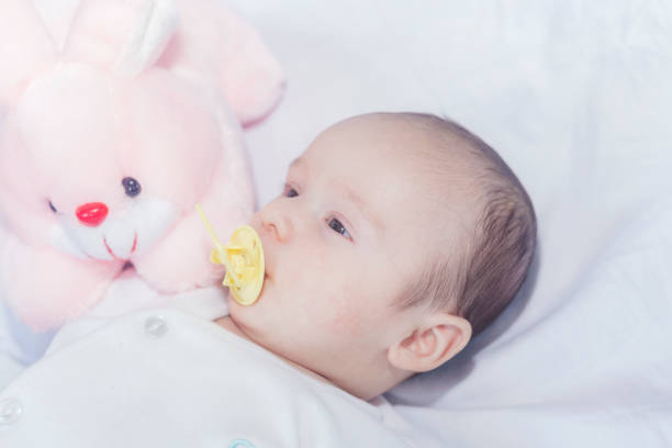 Baby sucking Pacifier looking at Camera. Child with Kids Smoother. Infant Boy Face Portrait with Dummy and Blue big Eyes in white Bed Room lying down on Stomach