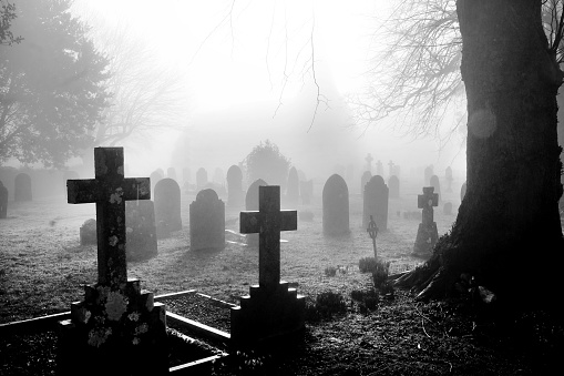 An english grave yard in the mist, in the foreground the head stones are in the shape of Christian crosses the head stones become more shrowded in mist and fog a distant dark shape of the church can be seen in the background, black and white photograph has a horror film feel about it.