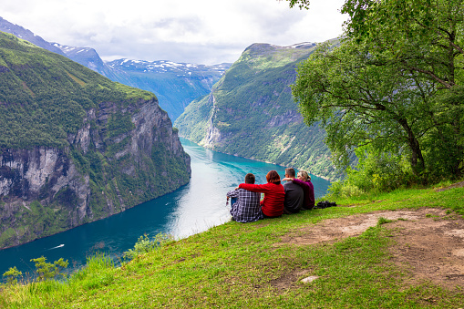 Two women and two men are sitting on the ground and looking at the majestic view in the Geirangerfjord on a summer day. On the both sides of the fjord, there are rocky mountains.