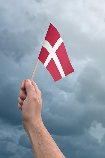 Hand holding Denmark flag high in the air, with a stormy, cloudy sky