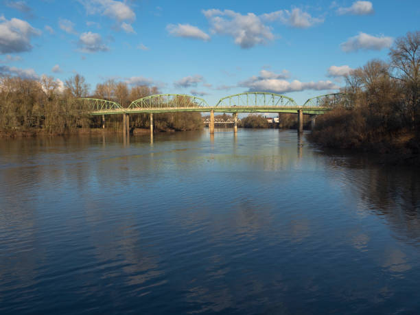 Bridges that span the Willamette River in Albany, Oregon. The most visible bridge is the Ellsworth Street Bridge. It carries eastbound traffic of Route 20. Behind it and only partly visible is the Lyon Street Bridge carrying westbound traffic. The Willamette River is shown with trees on the sides and a blue sky with puffy clouds. Divides Linn and Benton counties.