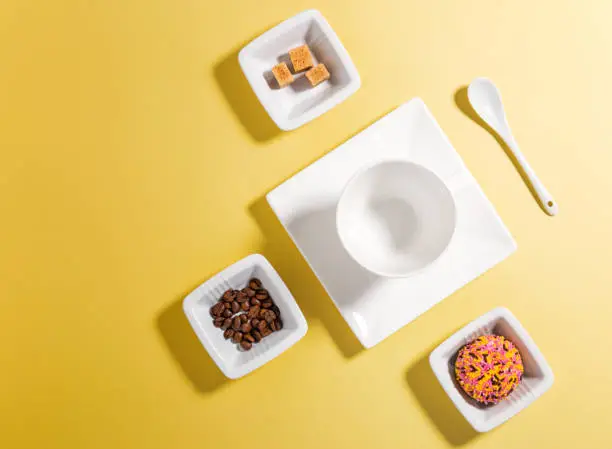 Top view of white porcelain with coffee beans, sugar and cake on yellow background