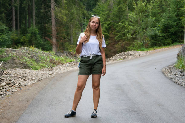 a beautiful girl is standing in the middle of a mountain road in a white t-shirt and shorts. - stripped shirt imagens e fotografias de stock