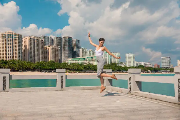 Photo of Cheerful and beautiful girl having fun on vacation, jumping on the pier on background of city. In China, Hainan city of Sanya. Town Sanya is popular tourist destination in China. View of island in bay
