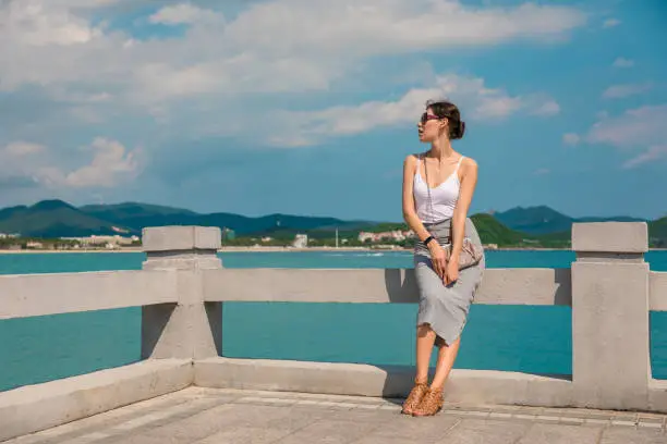 Photo of Cheerful and beautiful girl resting fun in vacation, on the pier on background of city. In China, Hainan city of Sanya. View of island in bay. Town Sanya is popular tourist destination in China