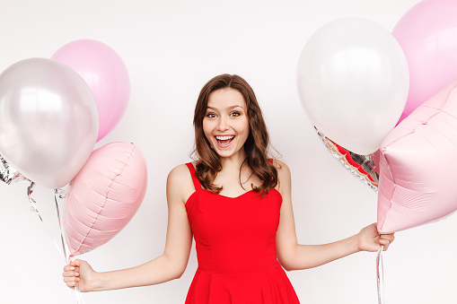 Pretty young woman in beautiful red dress laughing and looking at camera while holding two bunches of wonderful balloons and standing on white background