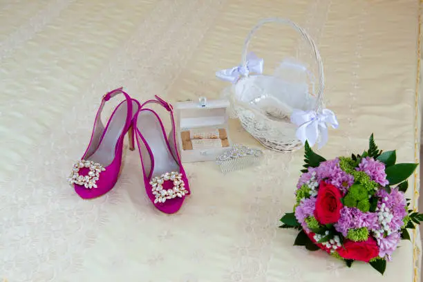 Still life on the double bed of the accessories of a bride on her wedding day, pink shoes with bright decorations, a wicker basket with the arras, a wooden box with the wedding rings, the bouquet of flowers and an ornament for hair.