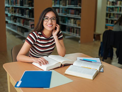 Female university student smiling looking at the camera while sitting in the library with a book.