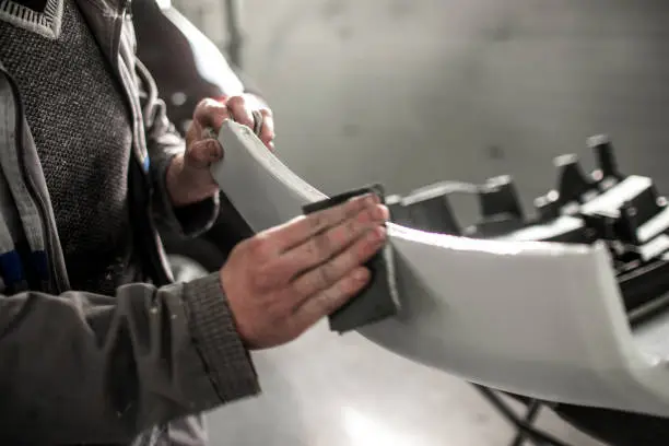 Body repair technician removing excess filling from an accident-damaged car bumper using a sanding paper.