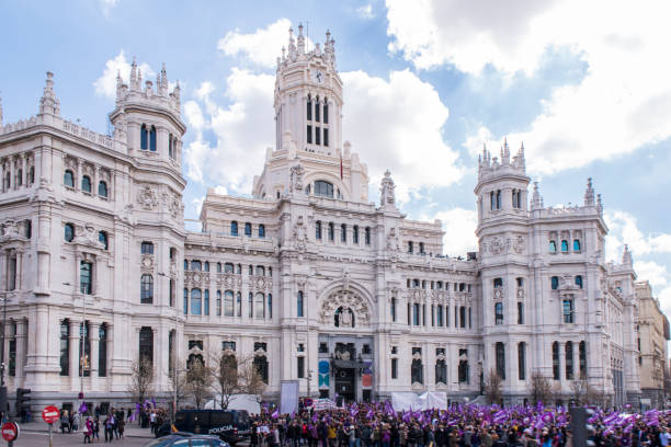 a crowd of people, most of them women wearing purple stuff, standing in front of the palacio de cibeles (cybele palace) in madrid city, spain. - spain flag built structure cloud imagens e fotografias de stock