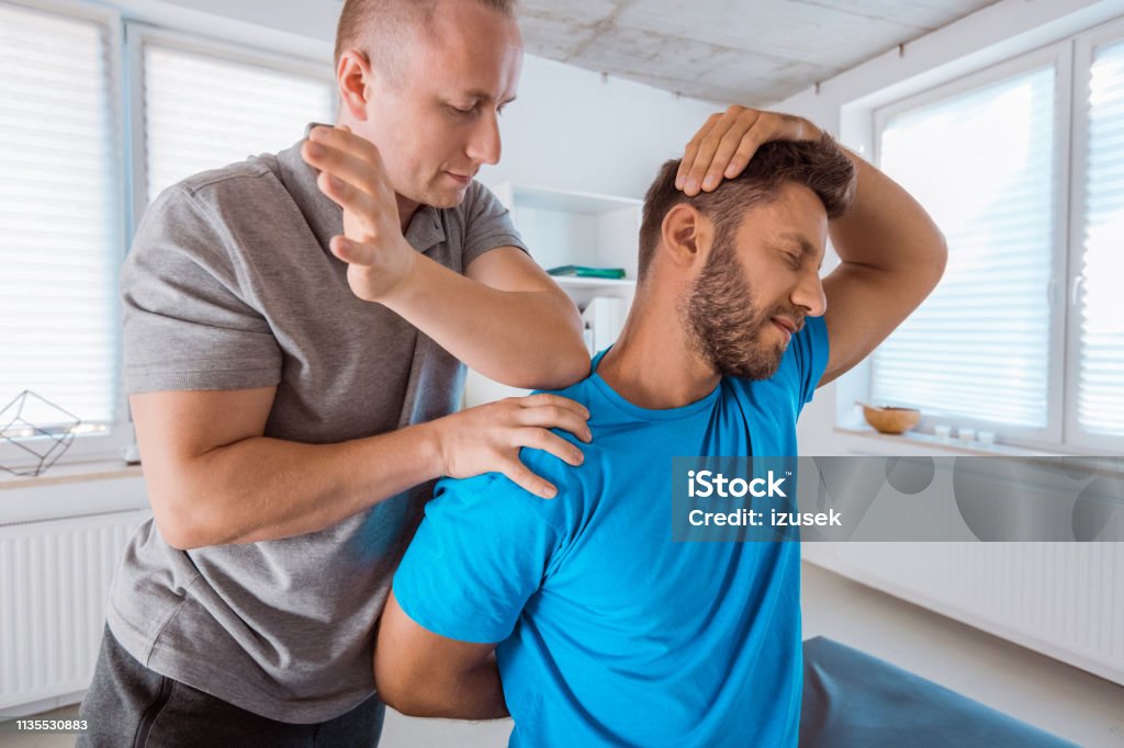 Physiotherapist massaging young man Physical therapist giving neck massage to young man. Patient sitting on massage table. Neck Stock Photo