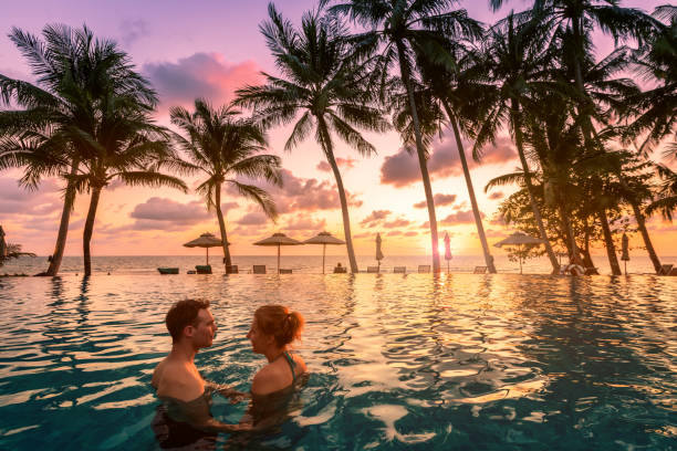 Photo of Couple at beach vacation holidays resort relaxing in swimming pool with scenic tropical landscape at sunset, romantic summer honeymoon island destination, coconut palm tree near the sea