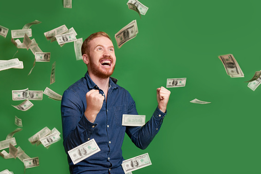 Excited emotional red haired man standing under rain of dollar bills
