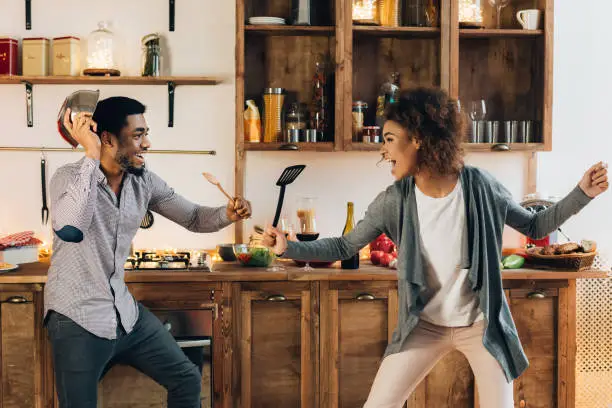 Funny african-american couple fighting with utensils tools, feeling playful in kitchen, copy space