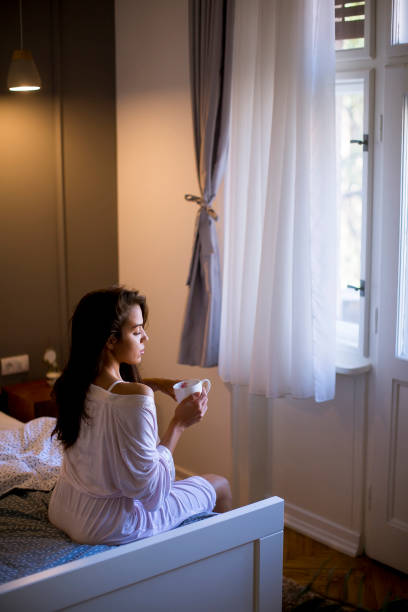 istock Young woman sitting on a bed with mug in bedroom 1135547634