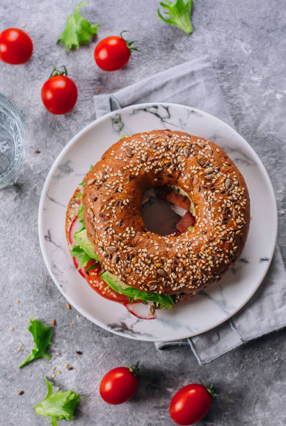 bagel con avocado, pomodori, ravanello e foglie verdi su piastra di marmo su sfondo tavolo in cemento. pranzo sano. delizioso concetto di cibo equilibrato. visualizzazione dall'alto - bun sandwich vegetable cream cheese foto e immagini stock