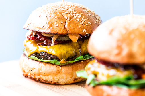 Close up image of two freshly flame grilled beef burgers loaded with melted cheese and crispy bacon with fresh green salad leaves, sandwiched between a toasted sesame seed bun. White backgroung with room for copy space.
