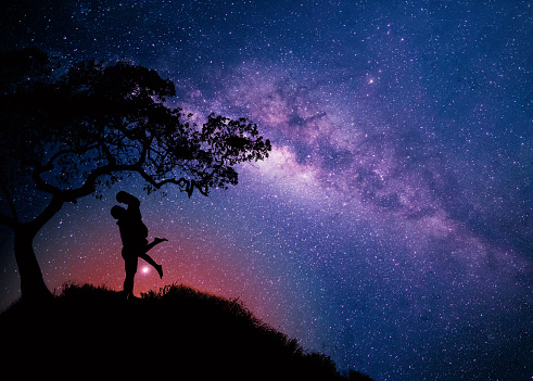 Milky Way with couple  under the tree on the hill. Landscape with night starry sky and silhouette of standing happy man and woman hugging in starry sky. Milky way with travelers,copy space.