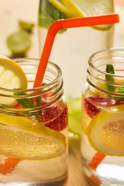 close-up of two glass jars illuminated by sunlight with refreshing cold lemonade water, lemon slices, red berries, mint leaves and drinking canes. Summer citrus soda background, vertical photo