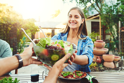 Shot of young woman taking a salad bowl from a friend while sitting around a table outdoors