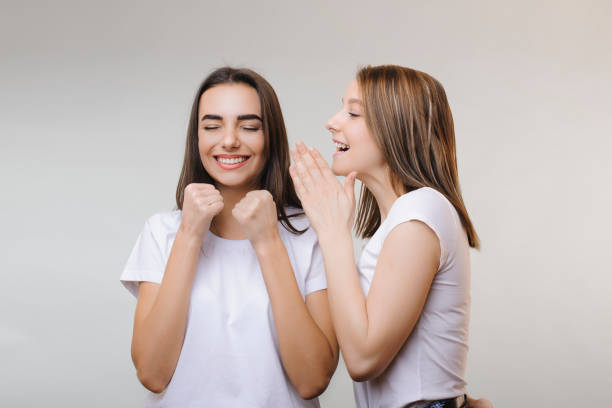 istock Two lovely women gossiping while one is telling something to another while another is laughing with closed eyes being happy against a grey wall.. Two women gossiping secrets 1135579719