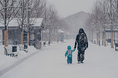 Outdoor shot of small child and father cover distance, being on way to home, hold hands, enjoy winter snowy weather. Back view of people walking on street during snowfall. Cold season concept