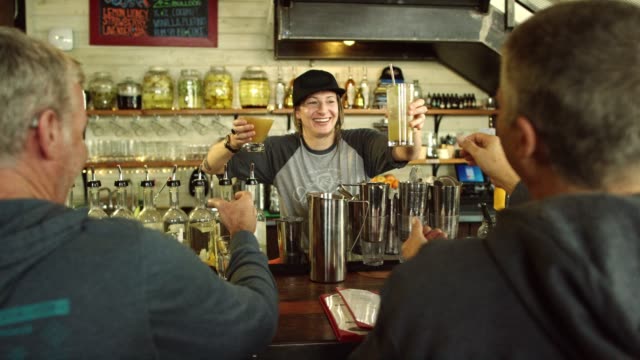 A Caucasian Female Bartender in Her Thirties Smiles at Hands Two Caucasian Men Their Cocktails while Working at a Bar