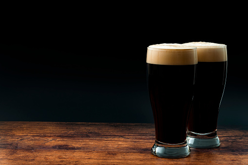 Alcohol abuse, bitter refreshing alcoholic beverage and dry stout concept theme with frothy glass pints of dark beer on wood table isolated on black background with copy space in bar or pub setting