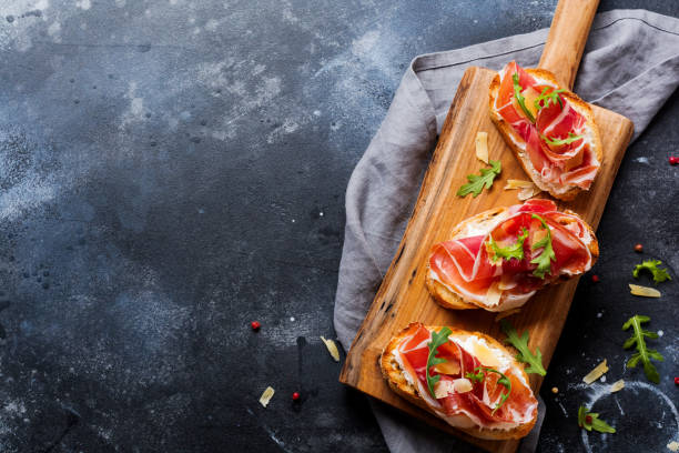 sándwiches abiertos con jamón, rúcula y queso duro en un soporte de madera sobre un fondo oscuro de hormigón viejo. estilo rustik. vista superior. - serrano chilli pepper meat ham spain fotografías e imágenes de stock