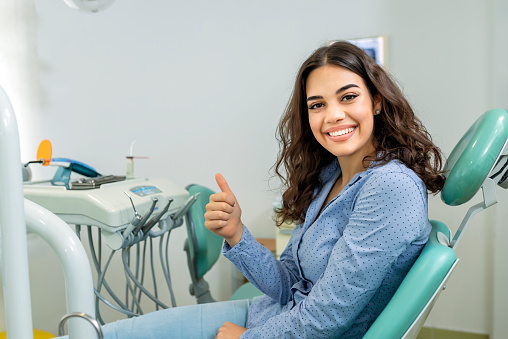Patient With Perfect White Teeth is Smiling After Dental Treatment in a Dentist Office With Medical Equipment in the Background. She is Satisfied with Visiting the Dentist. She is Showing Thumbs Up While Sitting in the Dentist's Chair.